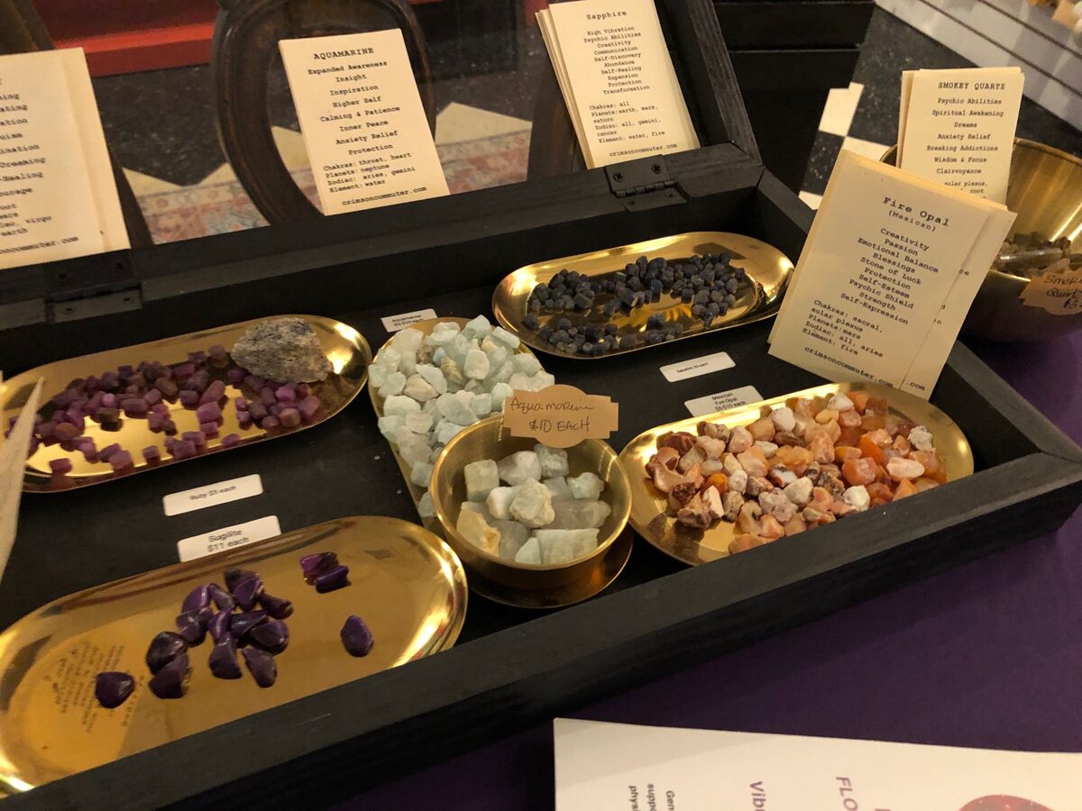 An open black display case on a purple cloth holding gold oval trays of loose tumbled stones in purple, pale blue, dark blue and orange, with printed property cards for aquamarine, sapphire, smokey quartz and fire opal standing behind them and handwritten price labels in front
