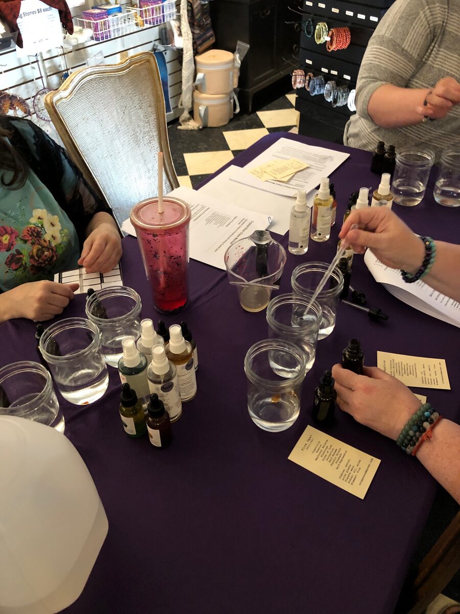 Hands working at a purple-clothed table set with small glass bowls, dropper bottles and printed notes during a flower essence workshop
