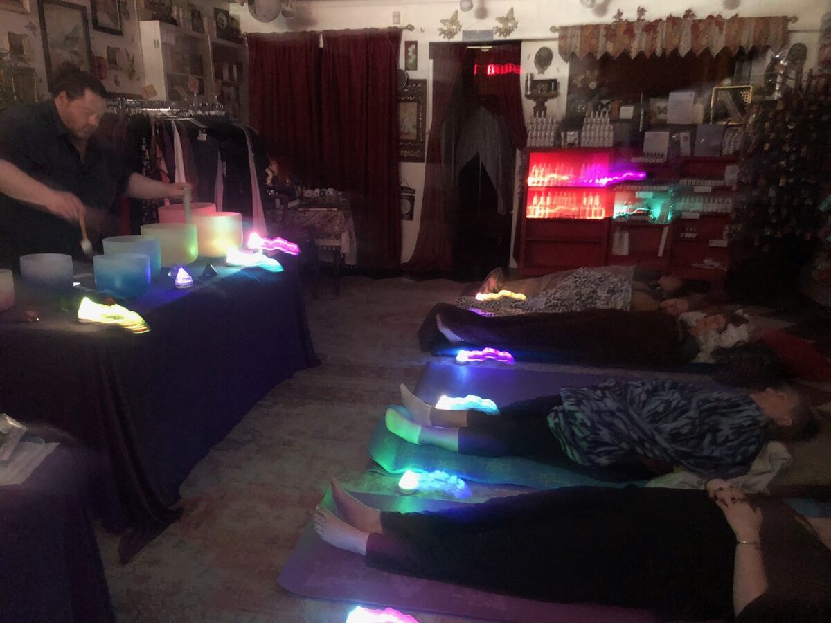 People lying on mats in a dim room during a sound bath, with coloured lights and a practitioner at a table of singing bowls