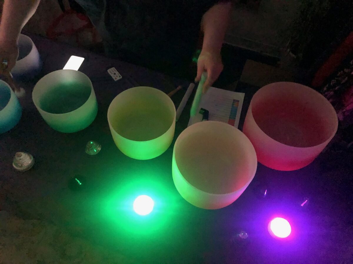 Five frosted crystal singing bowls on a dark surface lit green, yellow and pink by small LED lights, with two hands reaching in, one holding a pale mallet, beside a printed colour chart and a small remote control
