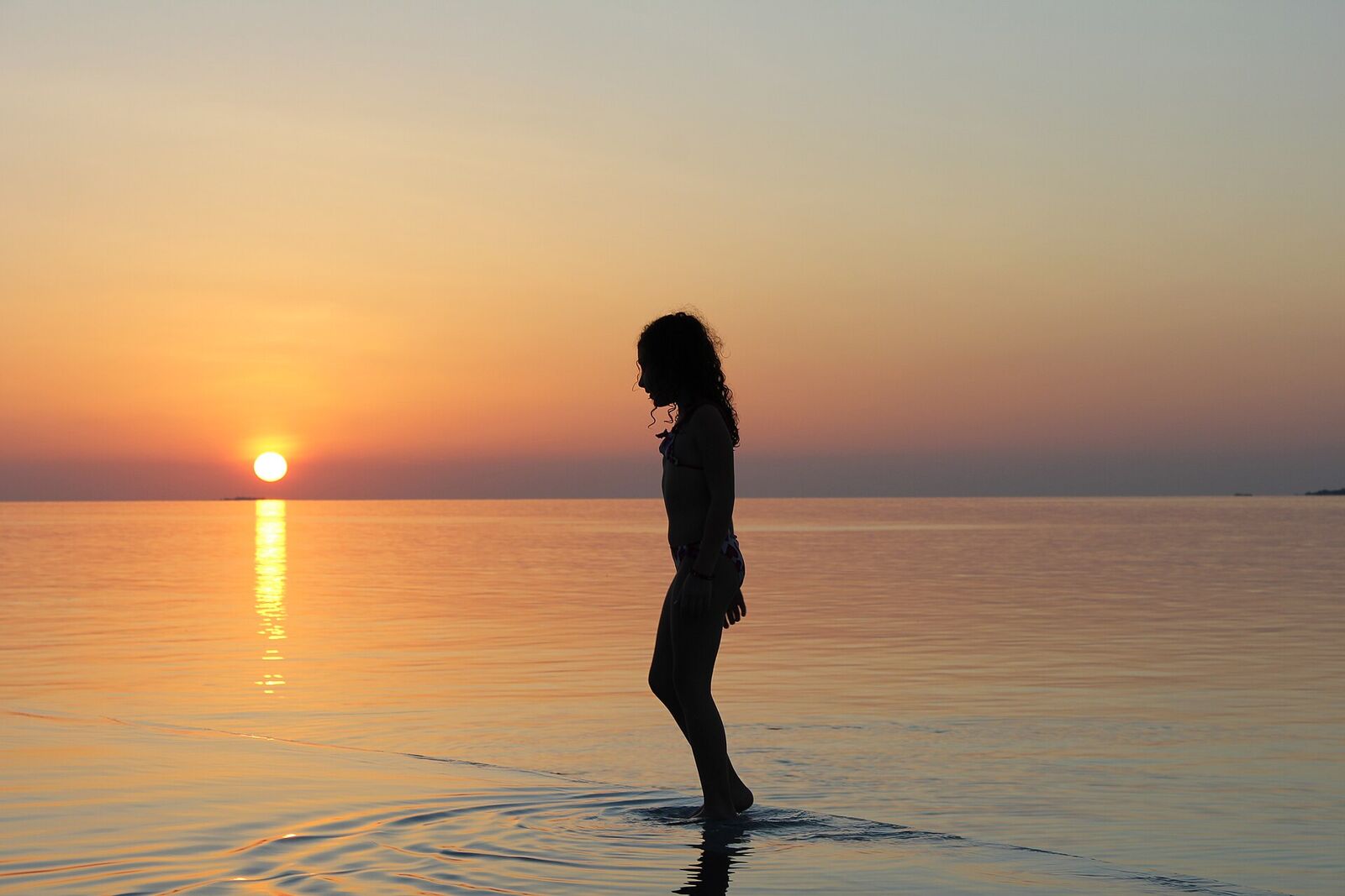 A person standing alone in shallow water at sunset, silhouetted against the low sun and its reflection