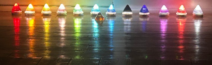 A row of small coloured lights in rainbow order reflecting on a dark glossy surface