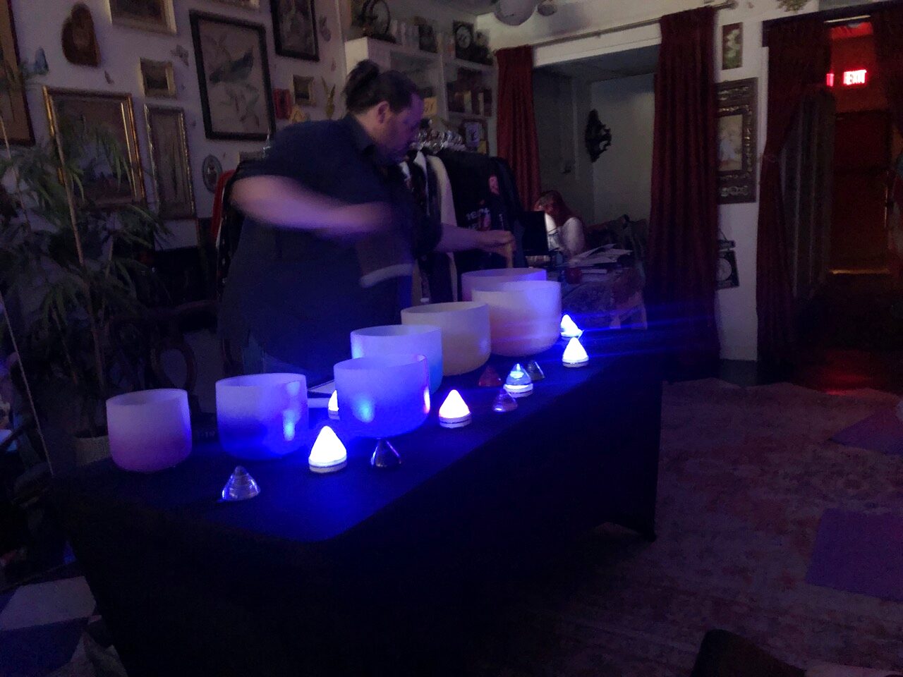 A man in a gray shirt leans over a row of six frosted crystal singing bowls on a navy draped table, each lit blue from a small cone light, in a dim shop room with framed pictures, red curtains and a clothing rack behind him