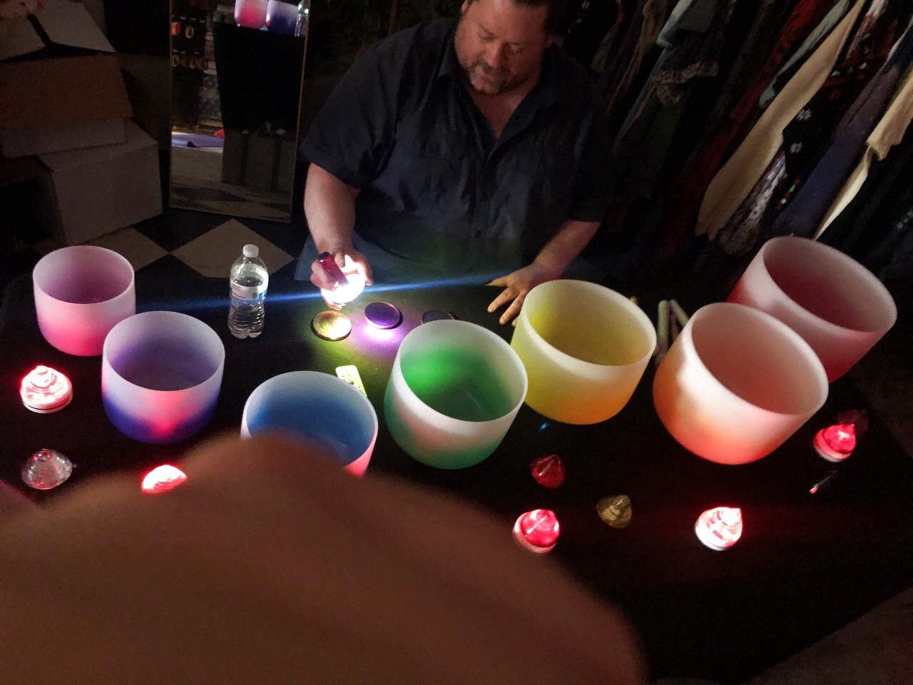 Coloured crystal singing bowls lit from within on a dark cloth, with candles and a practitioner seated behind them