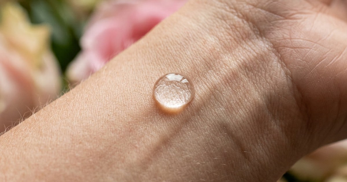 A single clear droplet resting on the back of a hand, with soft pink flowers blurred behind